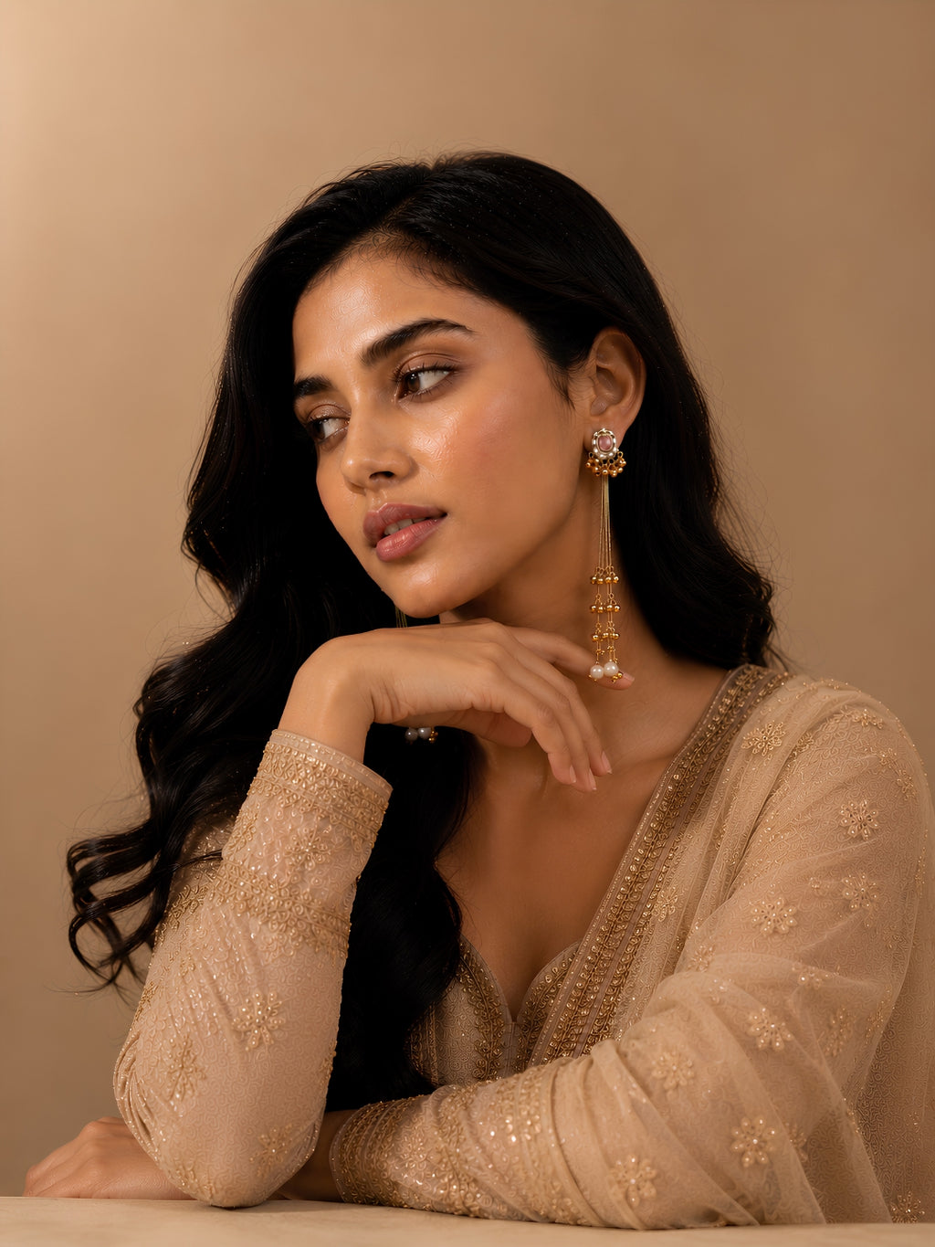 Woman wearing a beige embroidered outfit with gold jewelry against a beige background