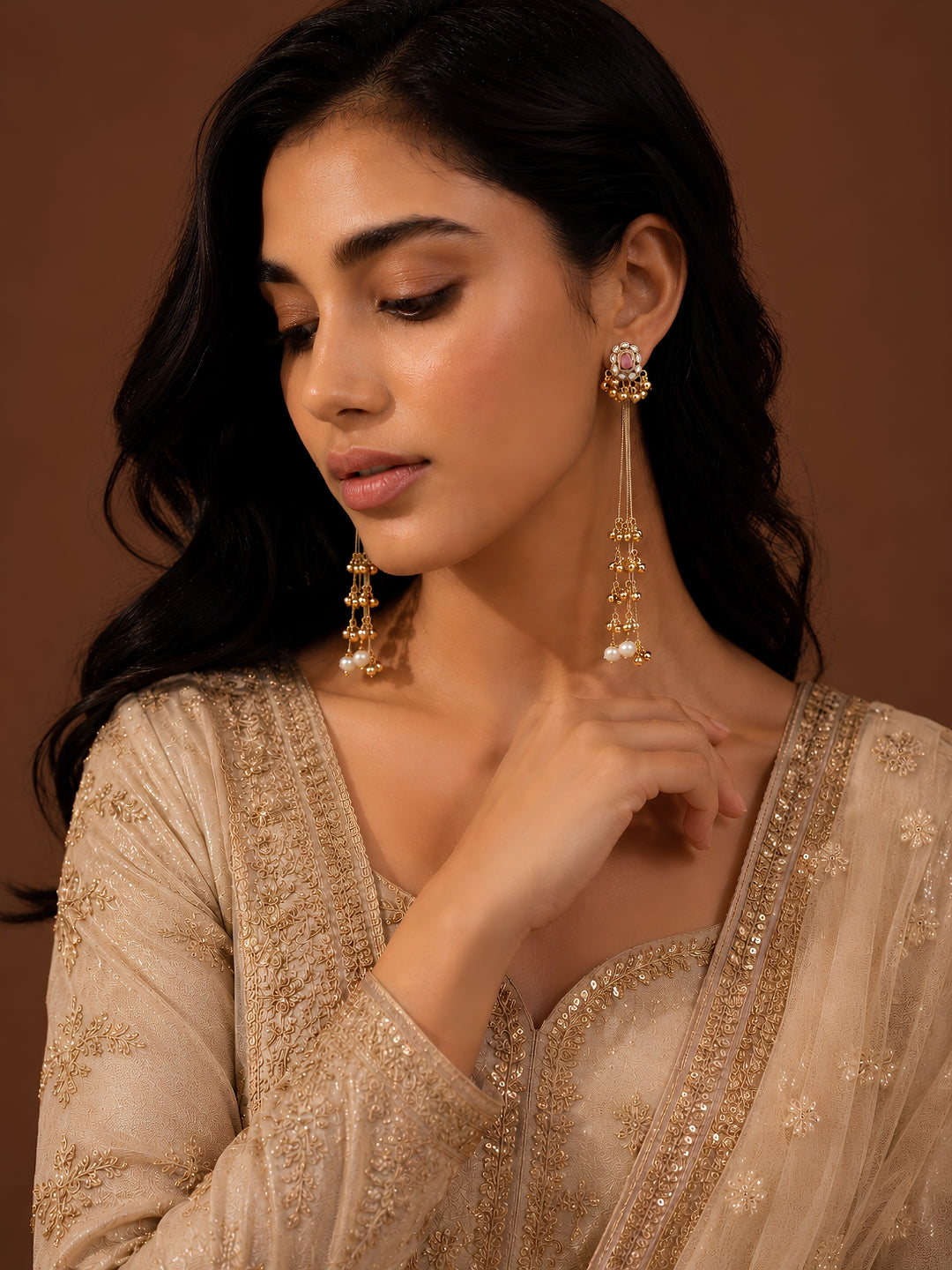 Woman wearing gold earrings and a beige embroidered outfit against a brown background