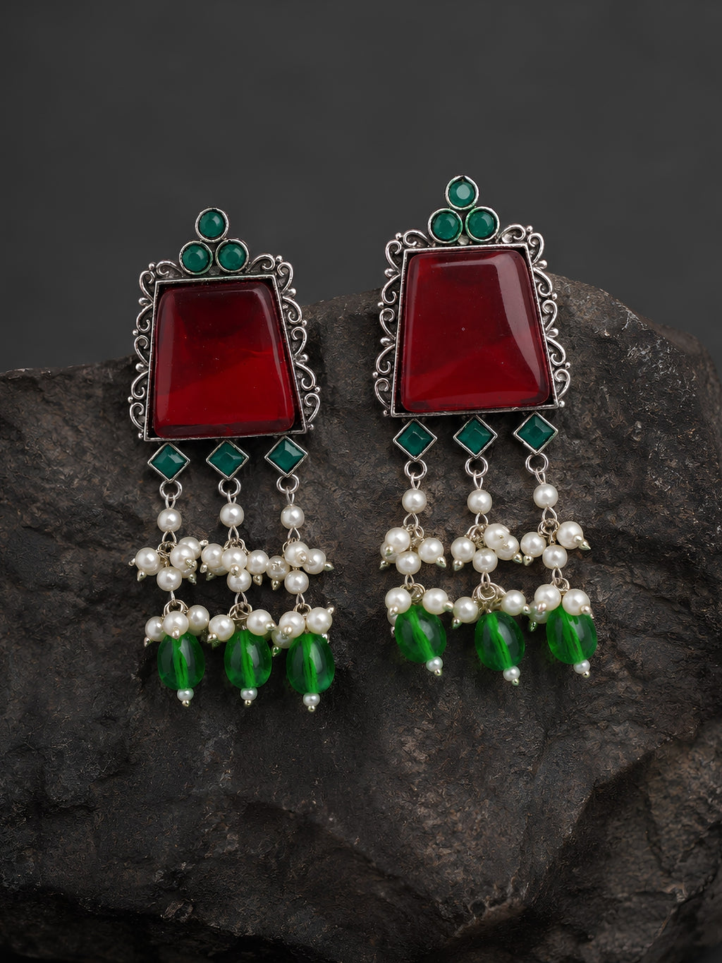 Pair of earrings with red stones, green beads, and pearl-like elements on a dark rock background.