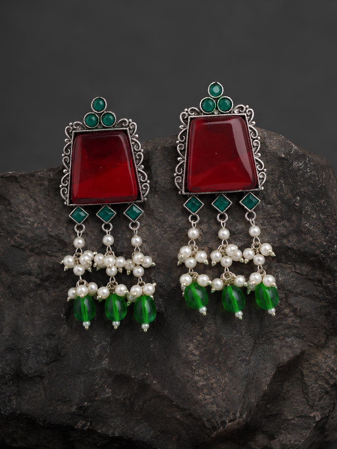 Pair of earrings with red stones, green beads, and pearl-like elements on a dark rock background.